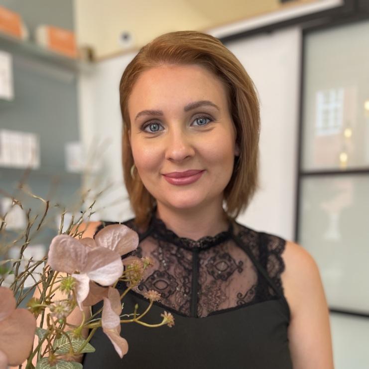 Fachkraft in schwarzer Arbeitskleidung steht lächelnd in einem hellen Behandlungsraum mit Produkten im Hintergrund.