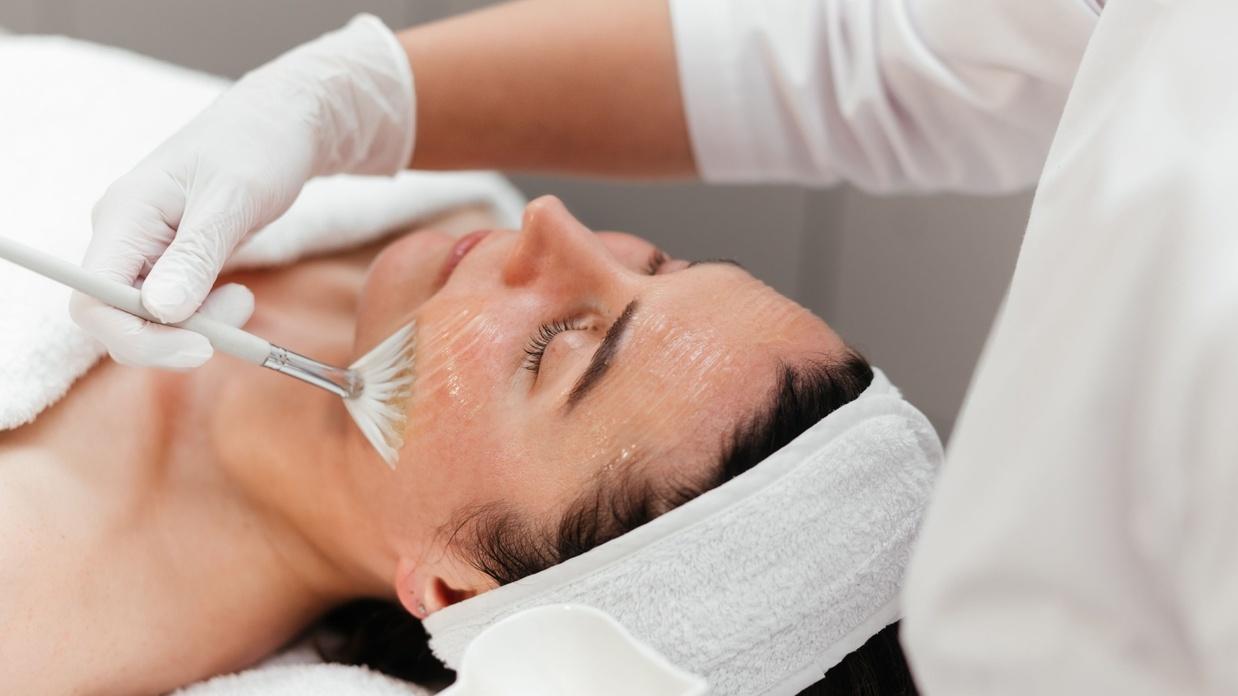 Frau erhält Gesichtsbehandlung mit einer Bürste auf der Liege; Creme und Handtuch im Hintergrund sichtbar.