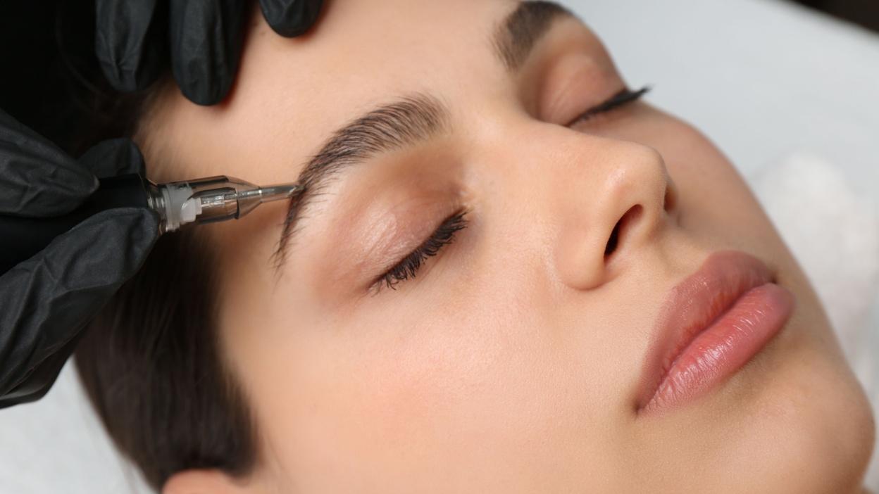 Nahaufnahme einer Person mit geschlossenen Augen, während ein Fachmann Microblading an den Augenbrauen durchführt.